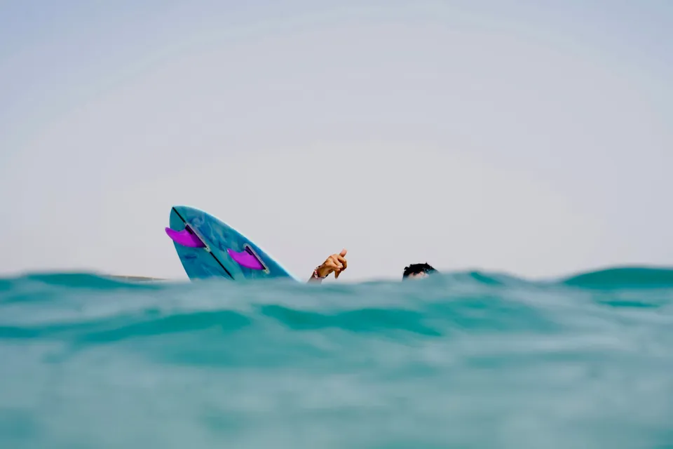Board nose and a shaka hand rising above turquoise water