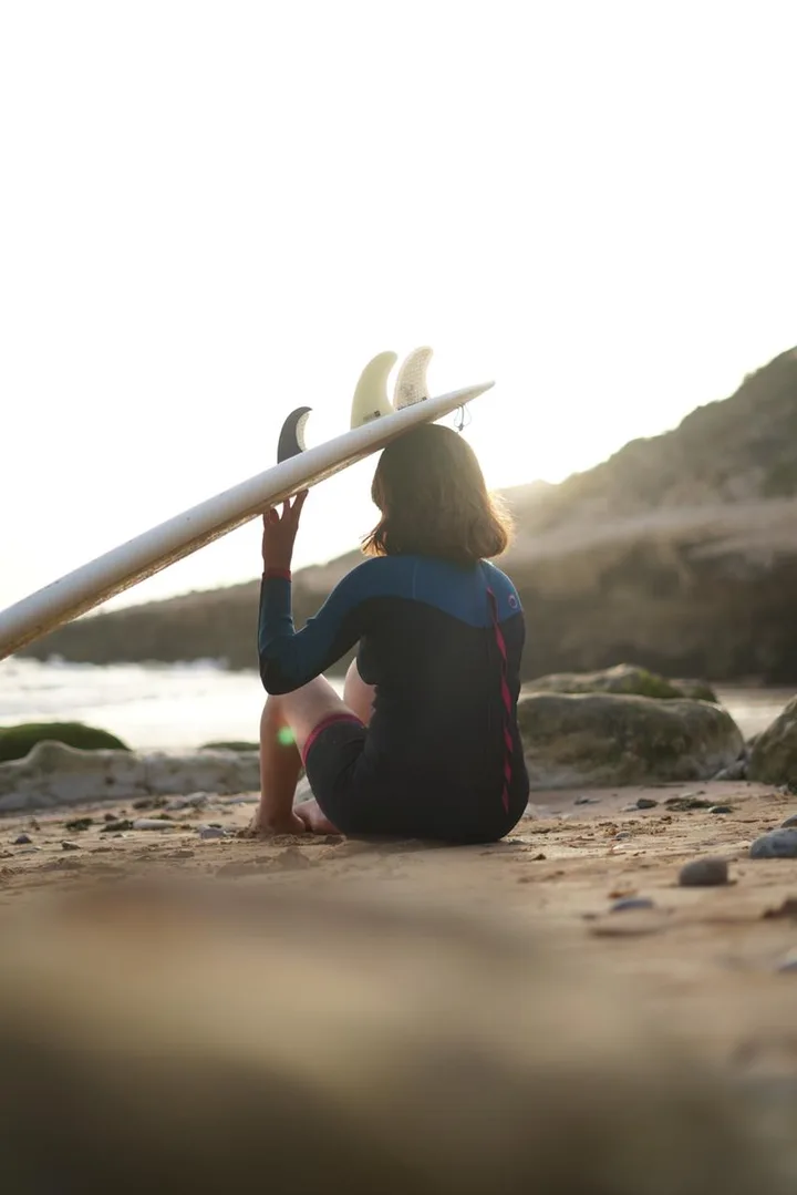 Surfer sitting on the shore with her back to the camera, board resting overhead, watching the ocean