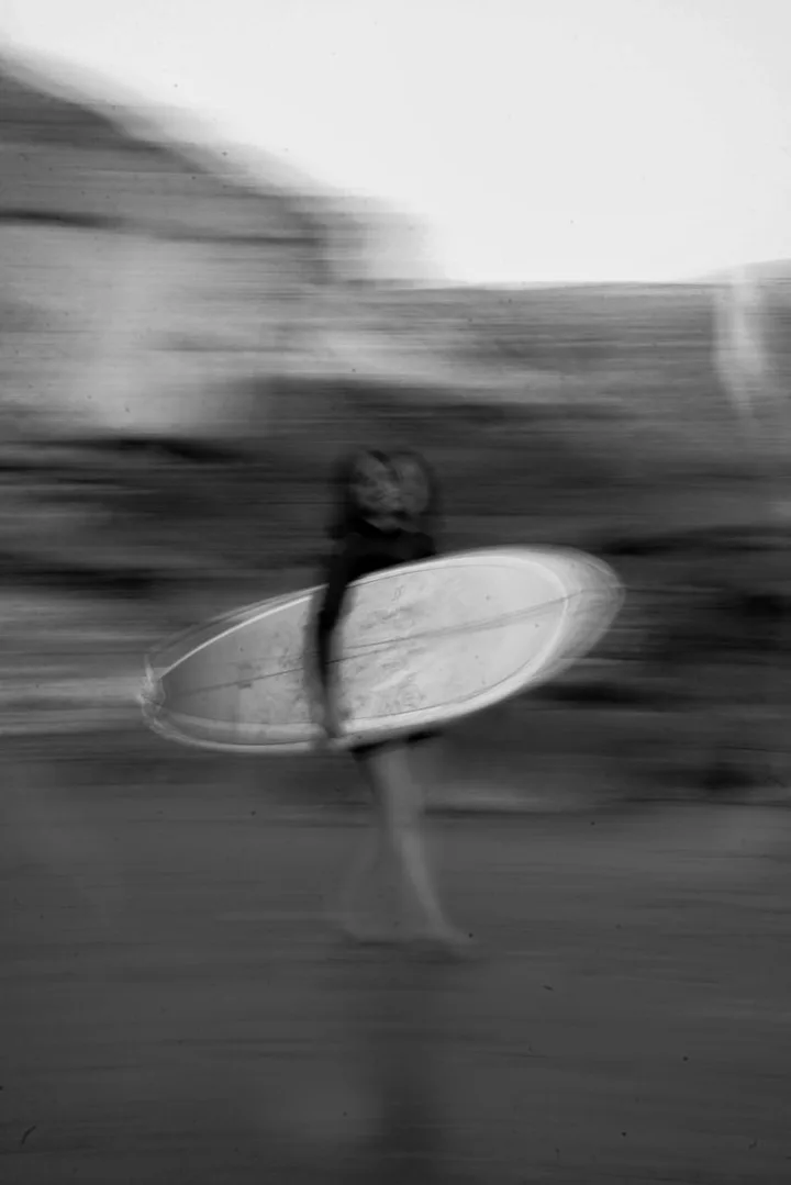 Black and white motion-blur shot of a surfer walking to the water with her board